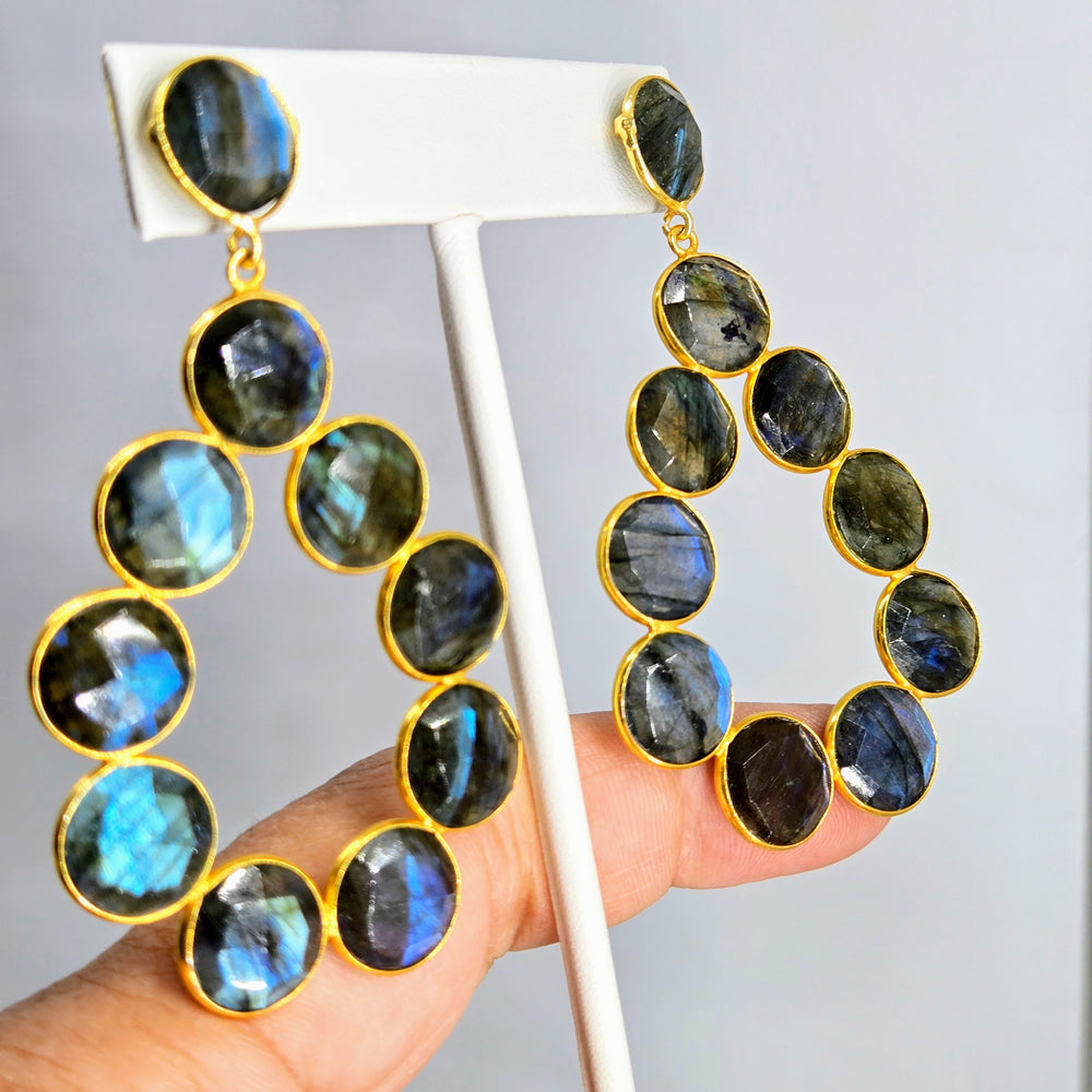A pair of hoop earrings with faceted labradorite stones set in 18k gold on a sterling silver base, displayed on a white stand.