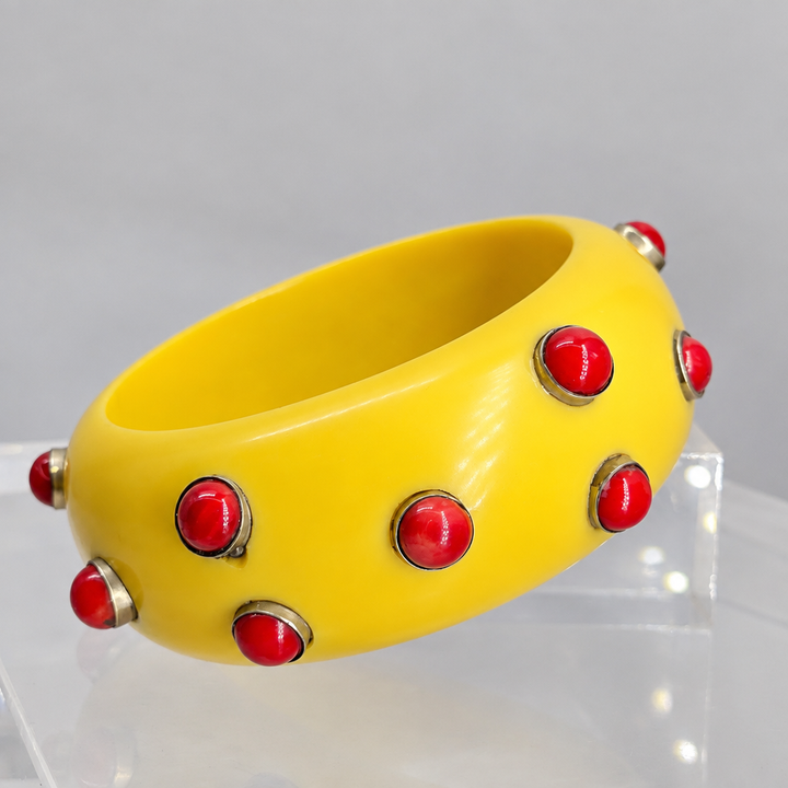 A bright yellow rigid bangle bracelet decorated with multiple red faceted gemstones set in silver-tone metal, displayed on a clear acrylic block against a neutral gray background.