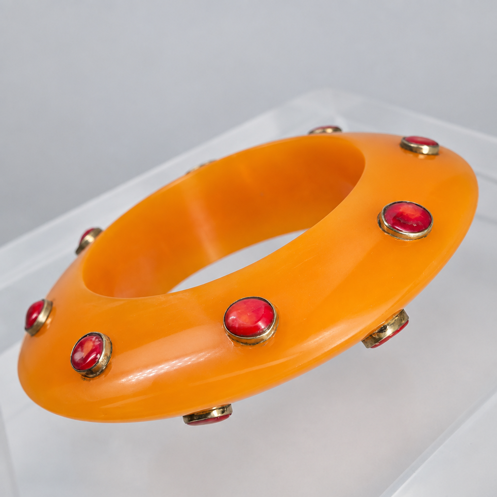 An orange translucent bangle bracelet decorated with multiple red cabochon stones set in gold-tone bezels, displayed on a clear acrylic stand against a neutral gray background.
