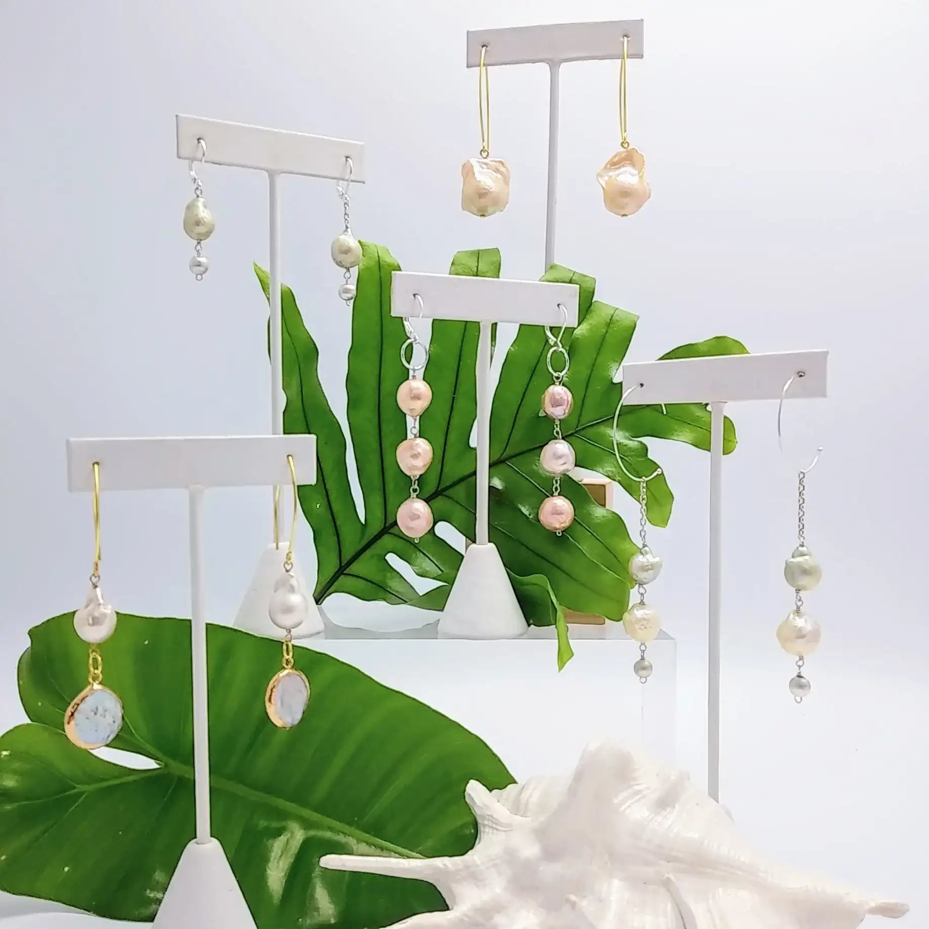 Delicate drop earrings featuring a cascade of iridescent pink and white pearls suspended from gold-toned hooks, arranged on minimalist display stands.